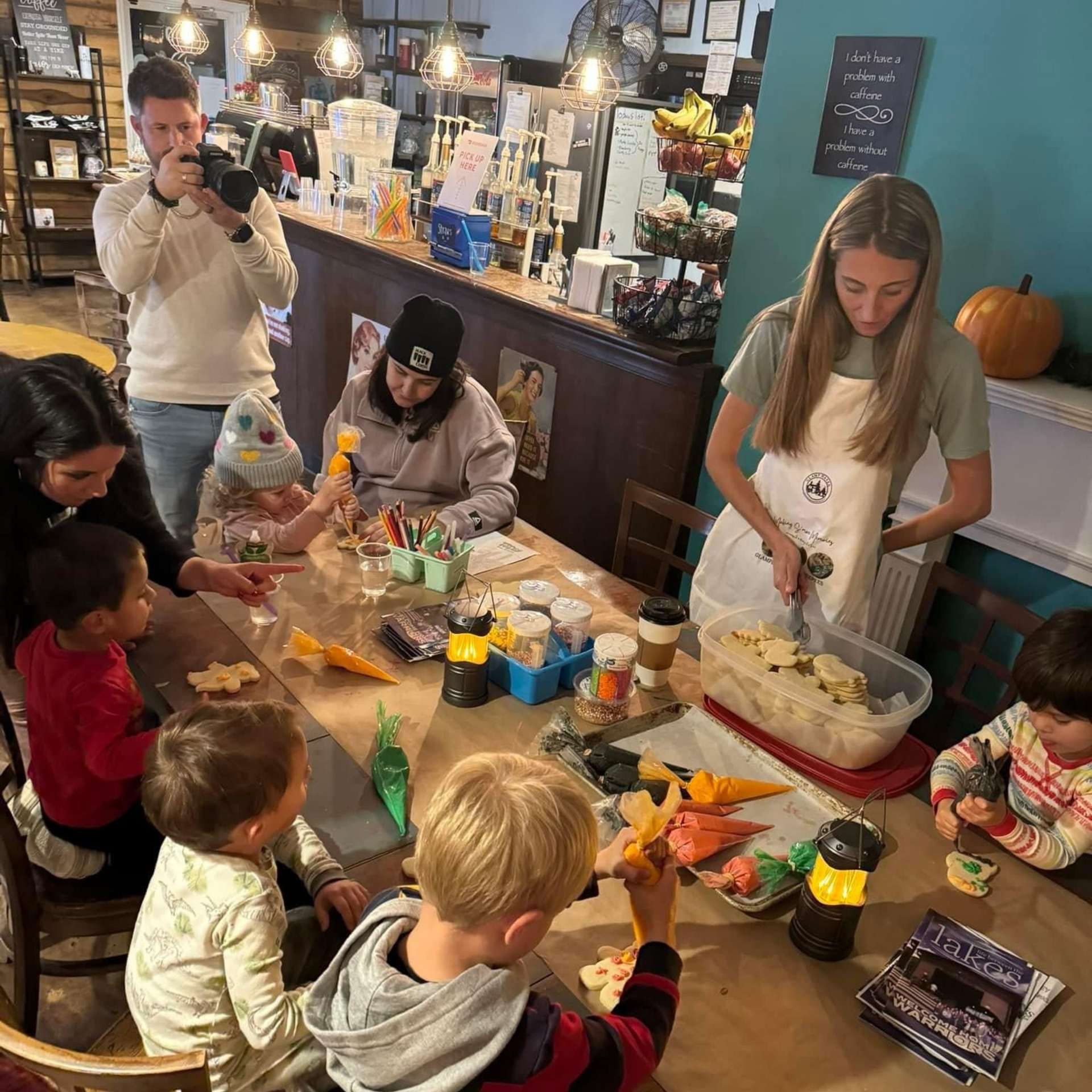 Children decorate cookies at a table. A woman cuts dough nearby while a man films. A pumpkin sits in the background.