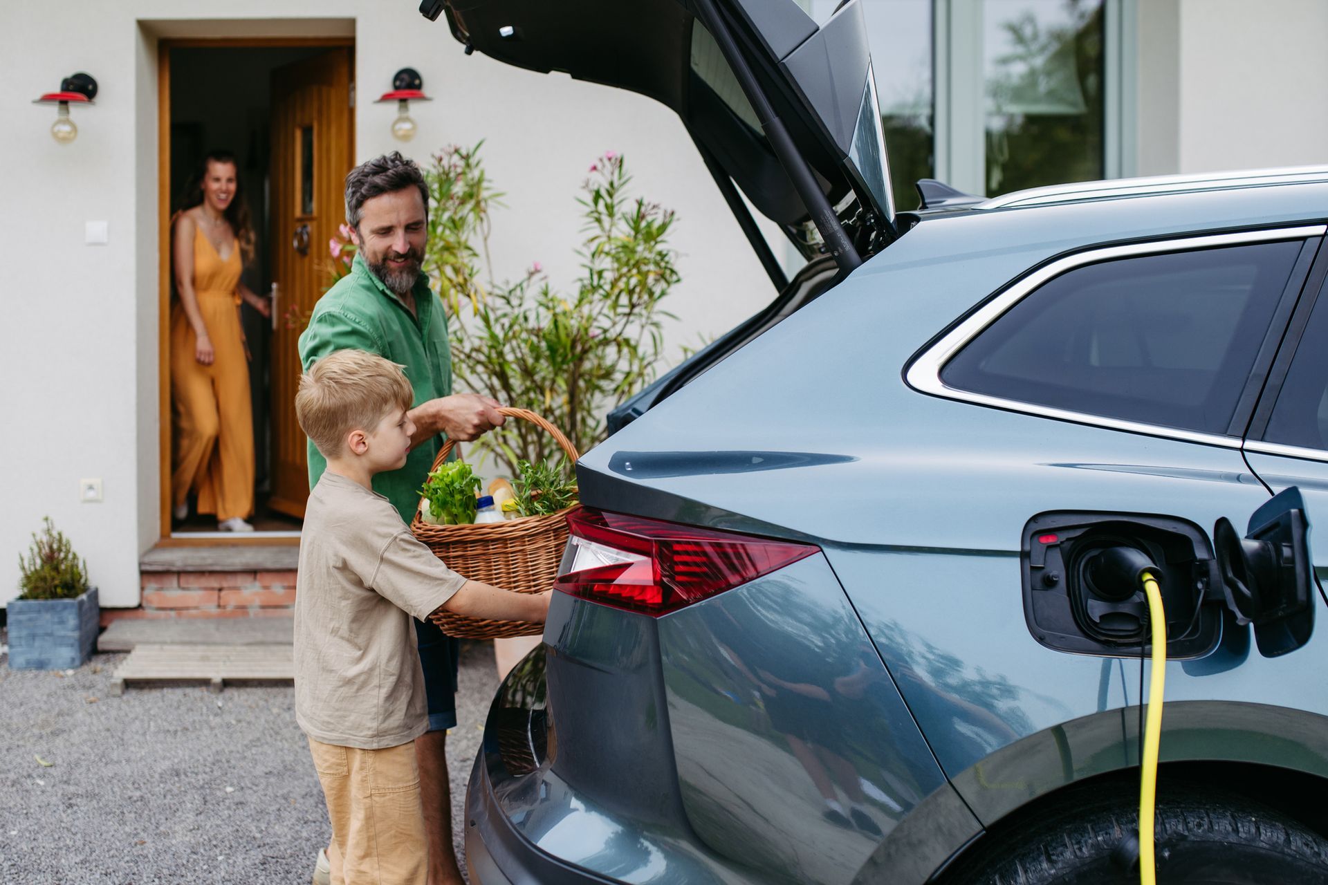 Electric car charging in a driveway while groceries are being loaded into the open trunk. Electric car charging in a driveway while groceries are being loaded into the open trunk.