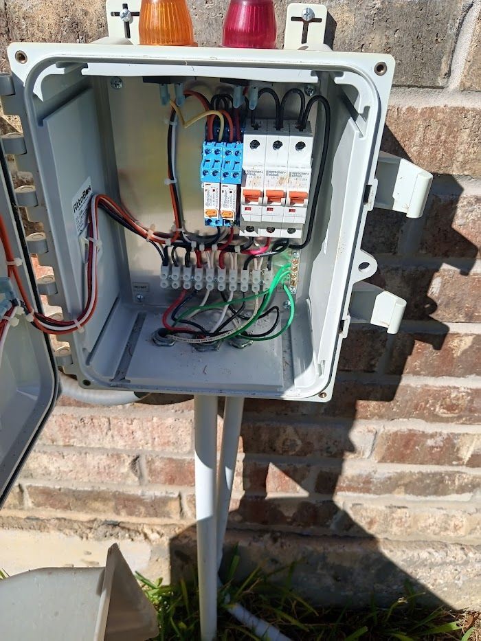 A box with a lot of wires inside of it is sitting on a pole next to a brick wall.