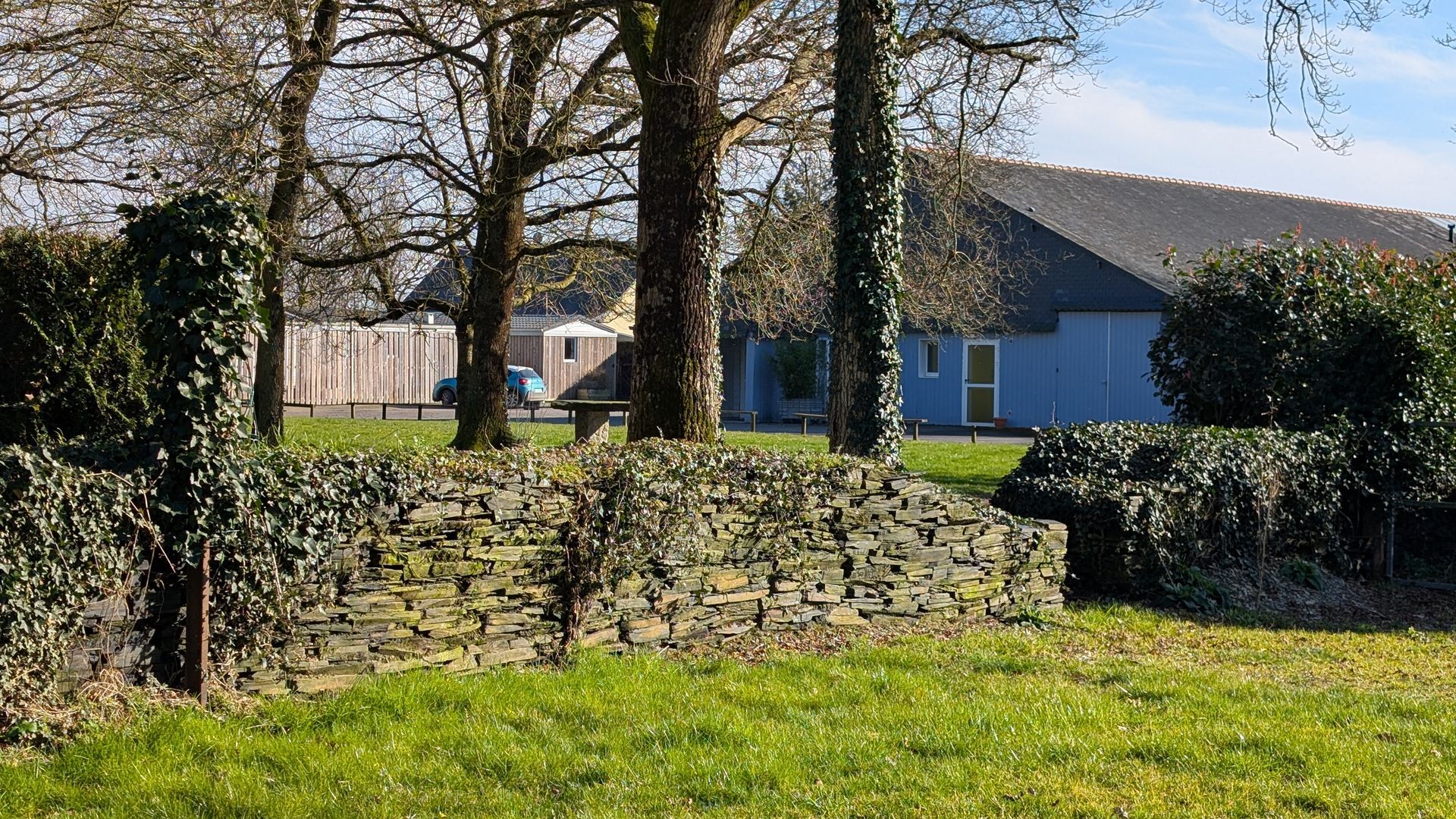 Un mur de pierre entoure un champ herbeux avec une maison bleue en arrière-plan.