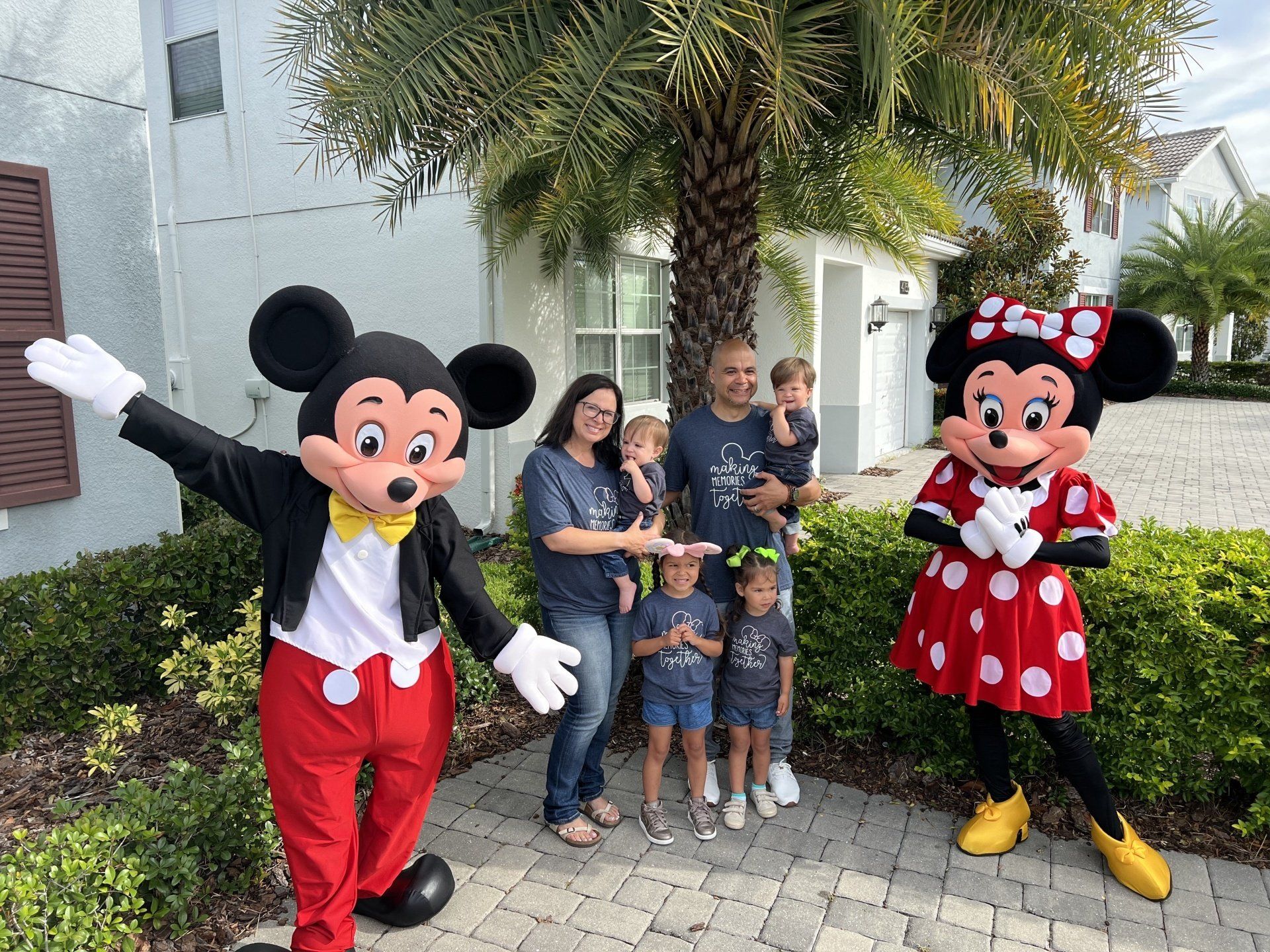 Family poses with Mickey and Minnie Mouse characters outdoors in front of a light-colored house, possibly on vacation.