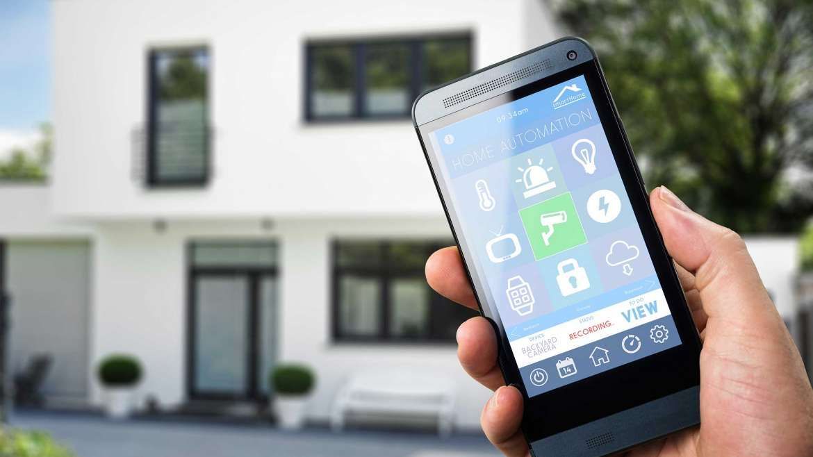 Hand holding smartphone with smart home app, house in background.