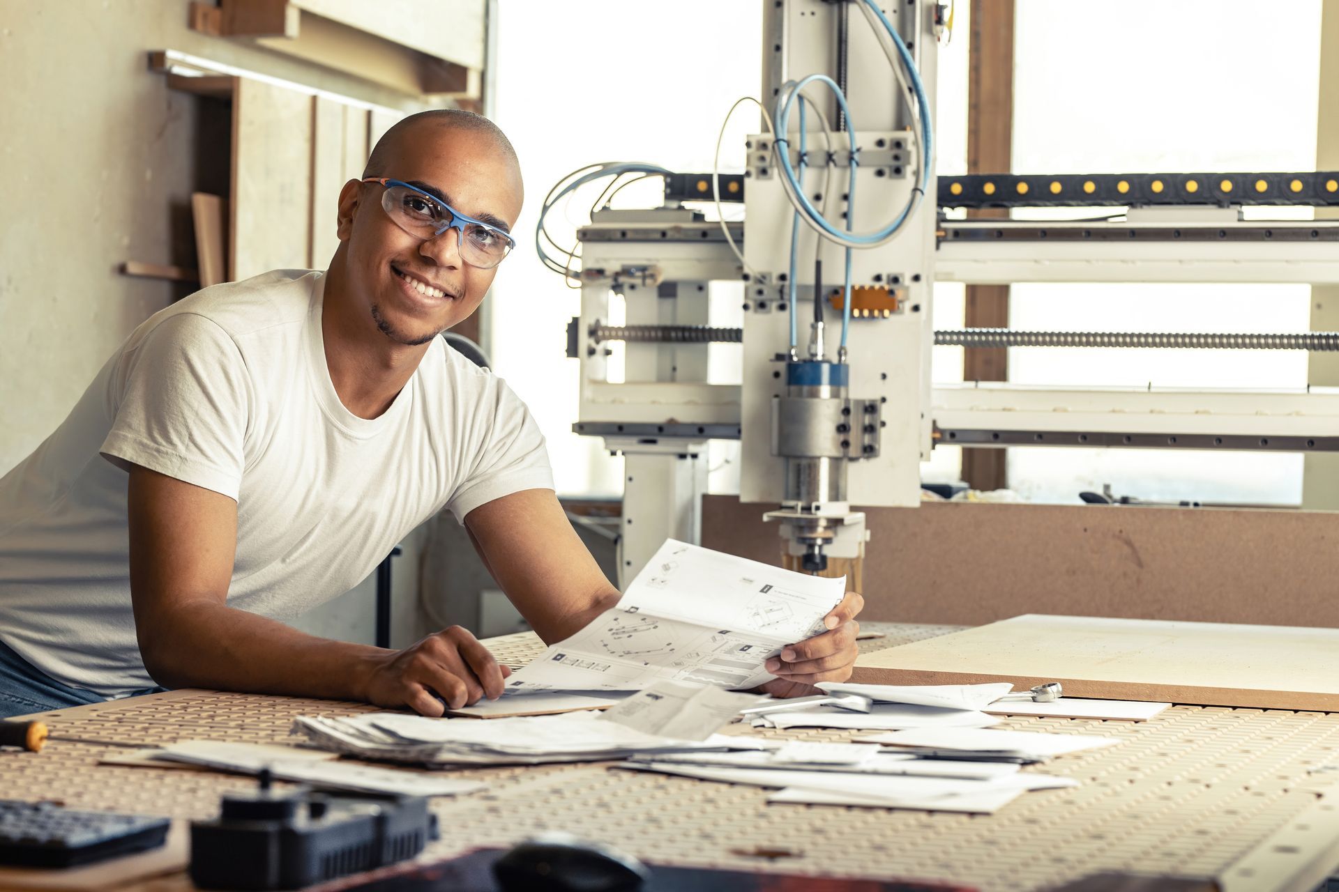Man in safety glasses smiles, holding blueprints at a CNC machine in a workshop.