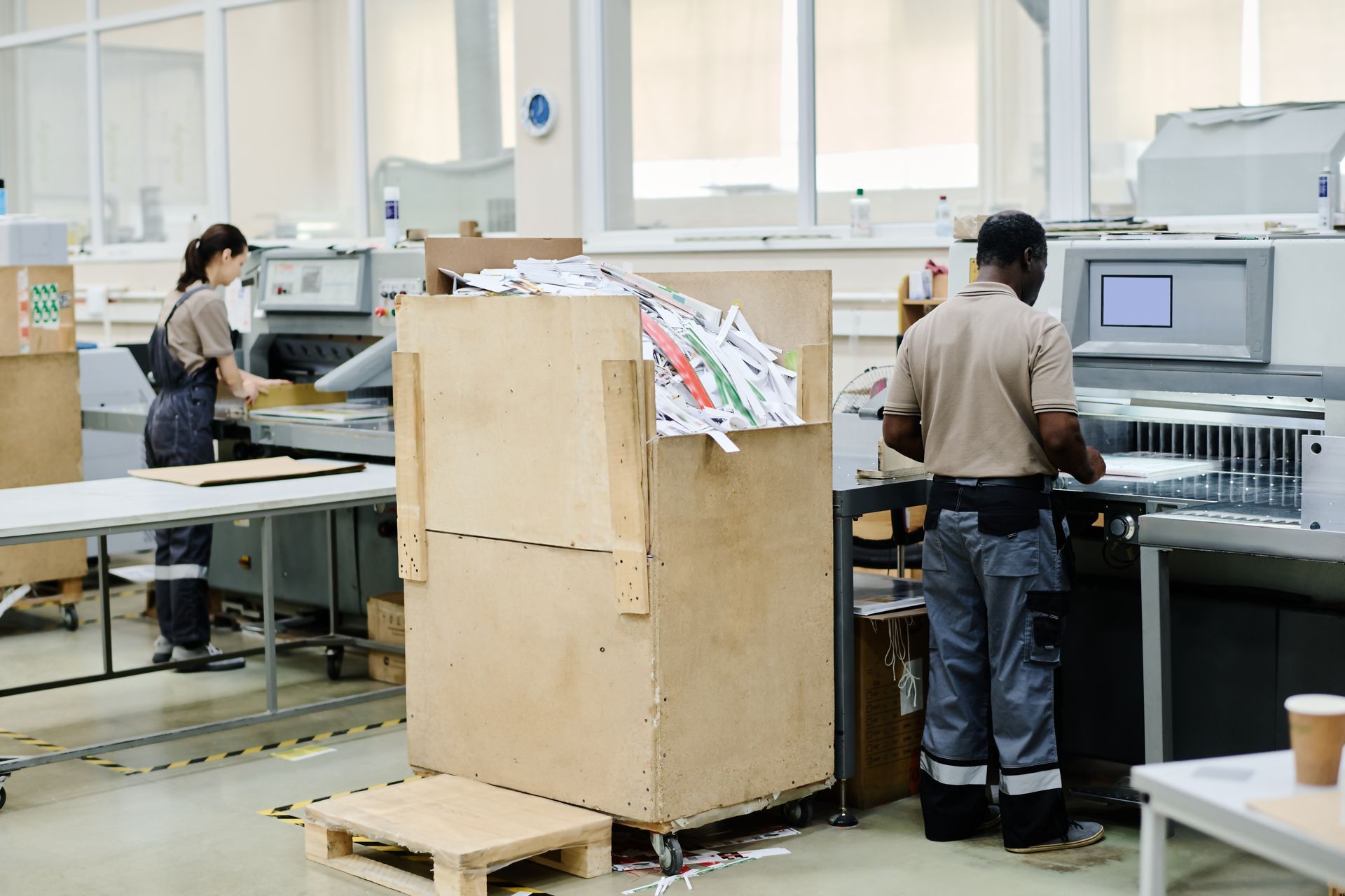 Two people working in a print shop, one at a machine, the other sorting paper. Wooden box in the foreground.