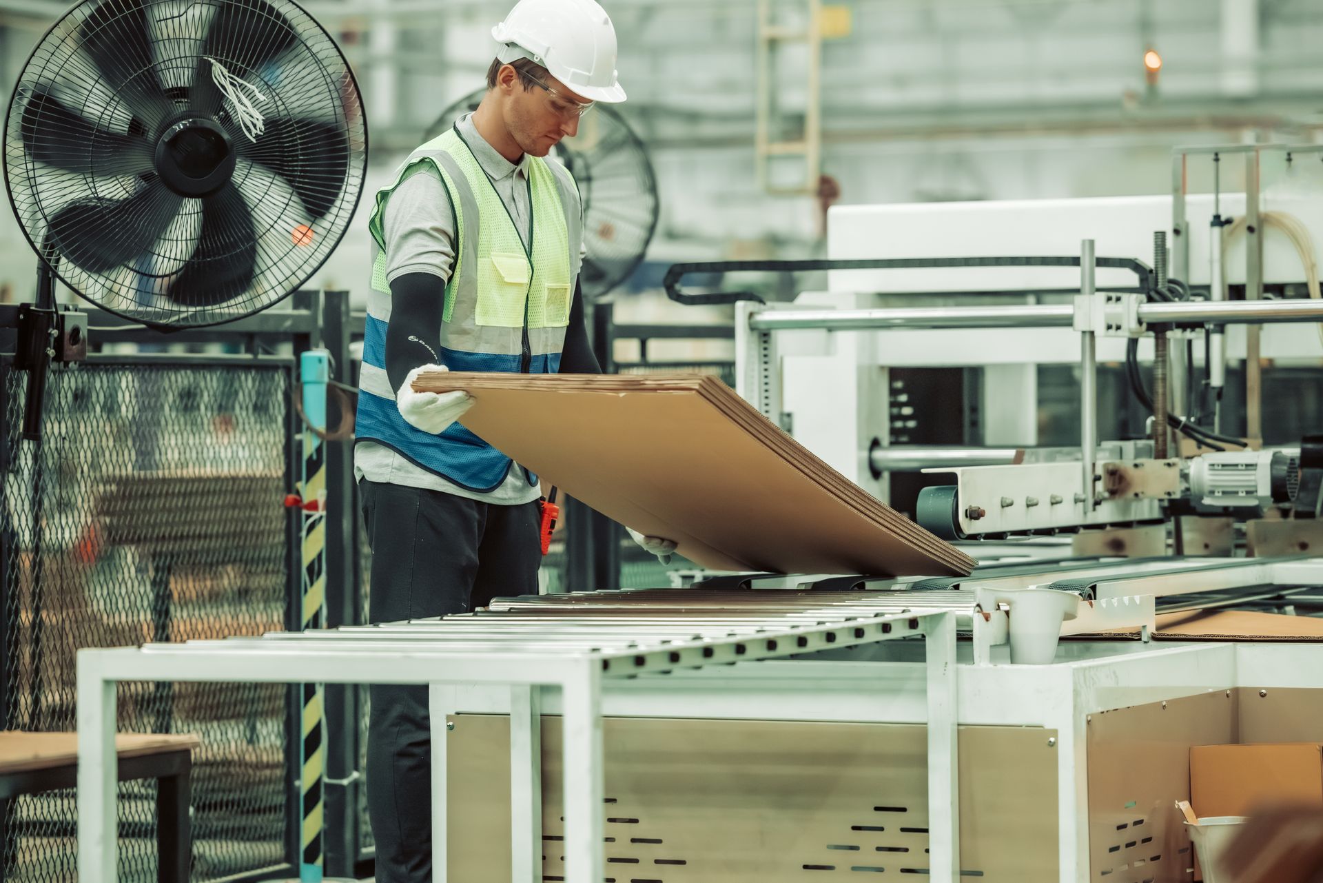 Worker in a factory wearing a hard hat and safety vest placing cardboard onto a machine.