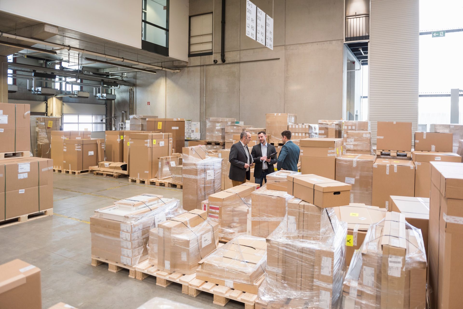 Warehouse interior with cardboard boxes on pallets. Three people in suits stand in the center.