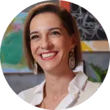 Woman smiles at the camera; wearing white shirt and drop earrings. Bright background with artwork.