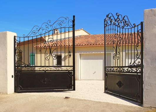 Open wrought-iron gate with black panels in front of a tan house and garage under a blue sky