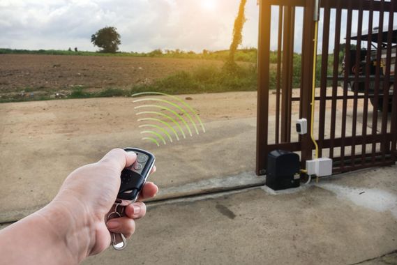Hand holding a remote opener toward a gated driveway at sunset, with fields in the background
