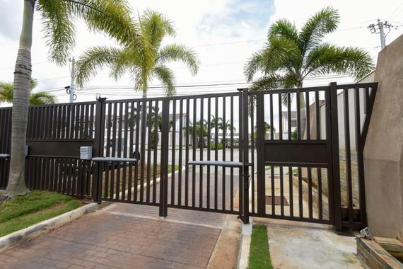 Black metal driveway gate with palm trees and a paved entrance under a cloudy sky