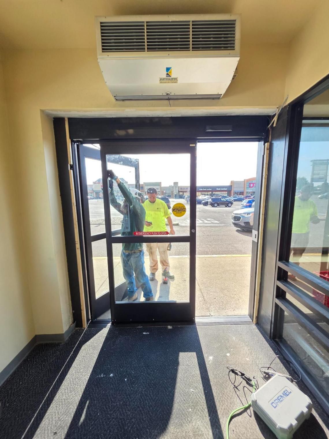 Indoor lobby facing open glass door, with two people outside and a white device on the floor.