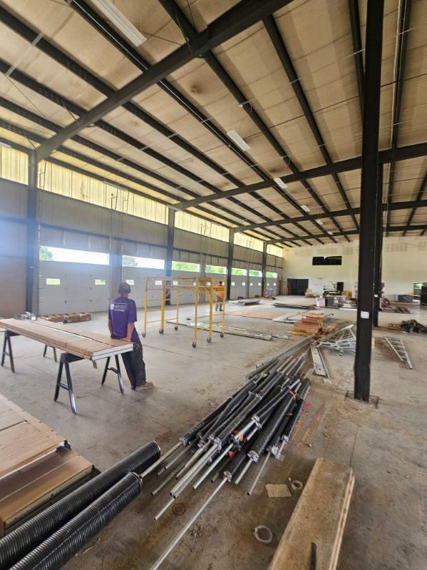 Inside a large unfinished warehouse, workers install framing near stacks of metal pipes and tools.