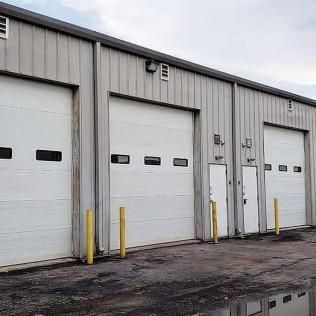 Row of white industrial garage doors on a gray warehouse exterior with yellow bollards and wet pavement