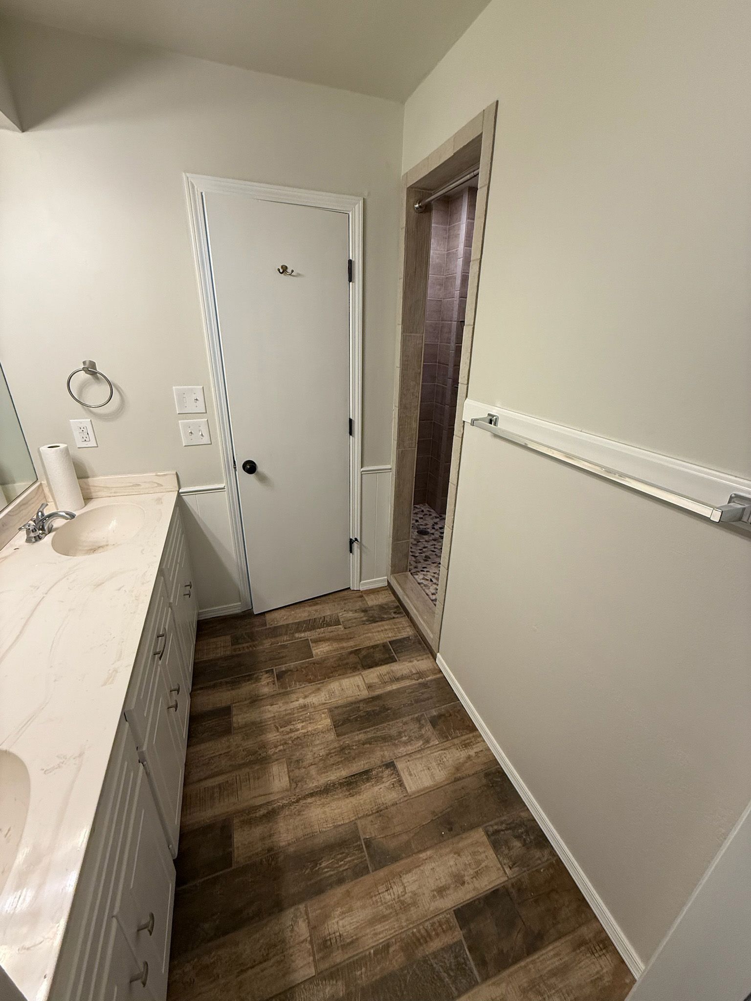 Bathroom with vanity, door, and shower, brown wood-look floor, and light walls.