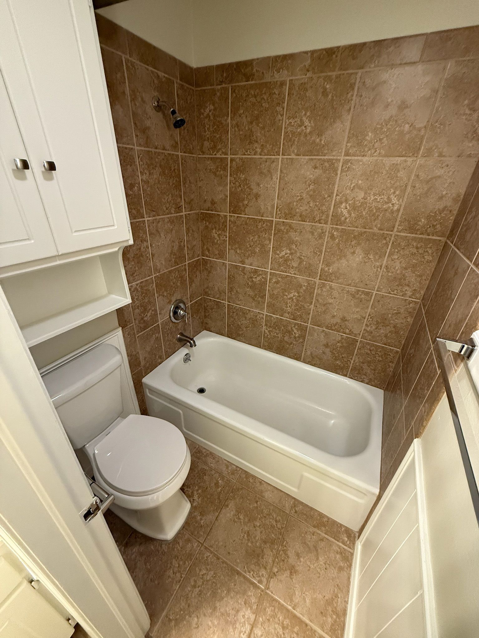Small bathroom with brown tile walls, white tub, toilet, and cabinets.