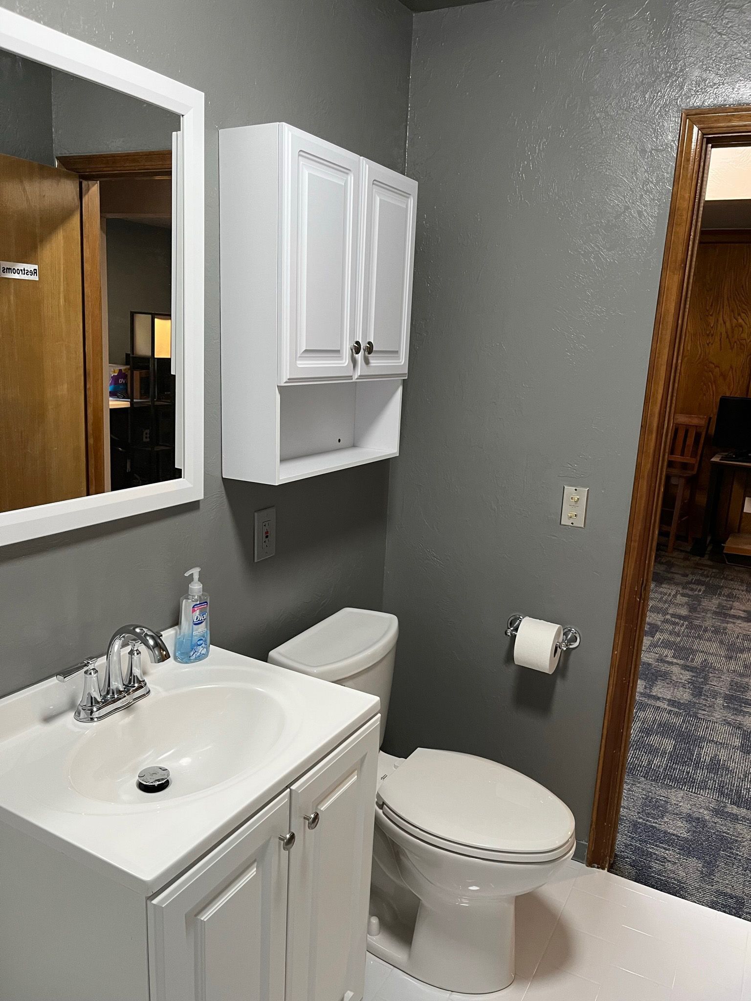 Bathroom with a white vanity, toilet, and cabinets. Gray walls and a doorway to another room.