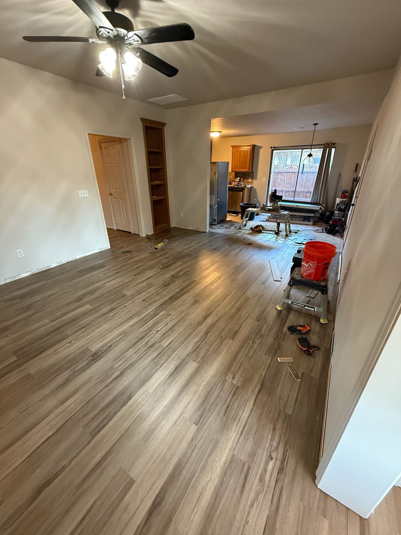 Room with newly installed wood-look flooring, open to a kitchen area. Red bucket and tools scattered about.