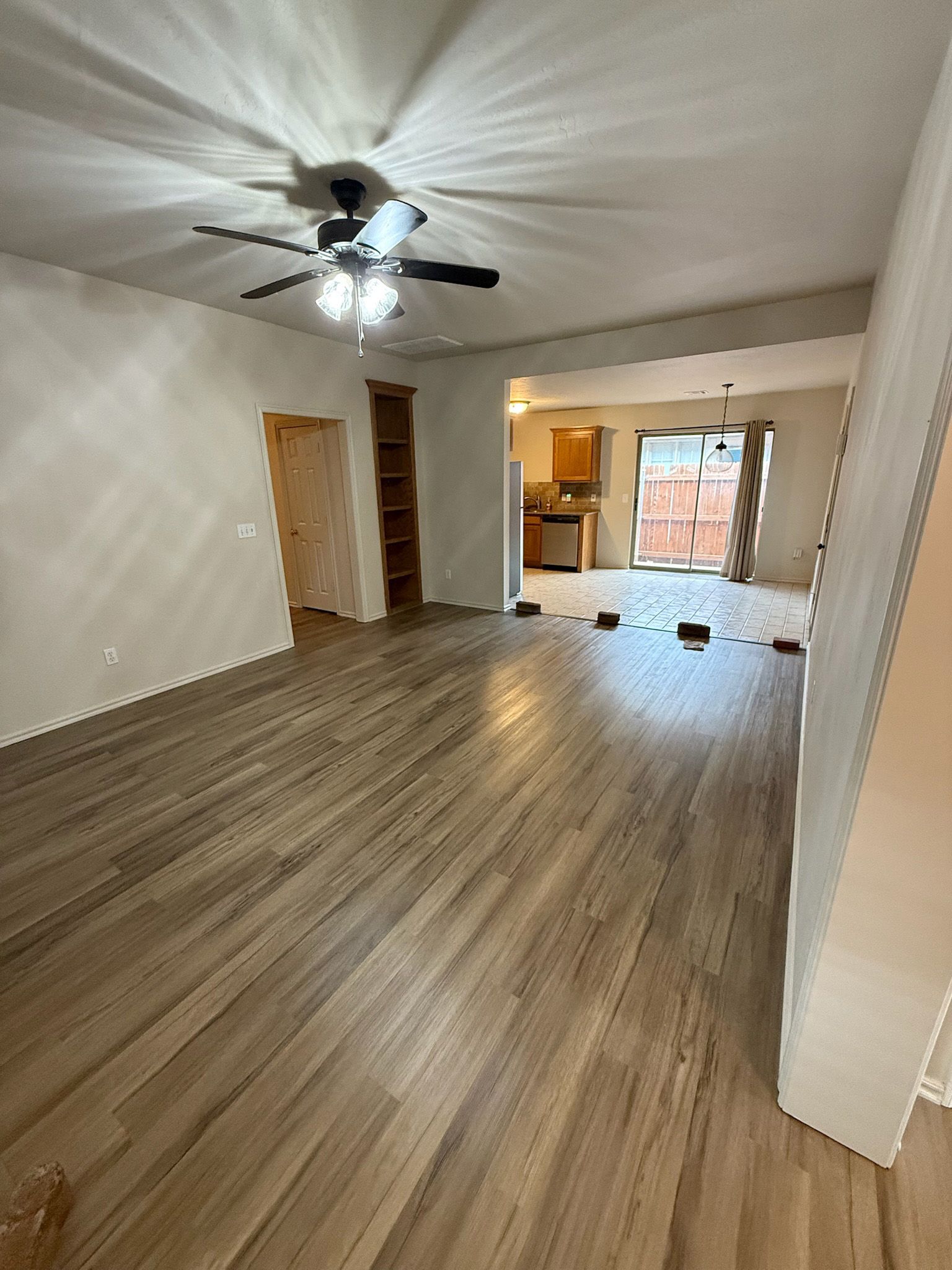 Open living room with light wood floors, neutral walls, ceiling fan, and a view into the kitchen and outdoor area.