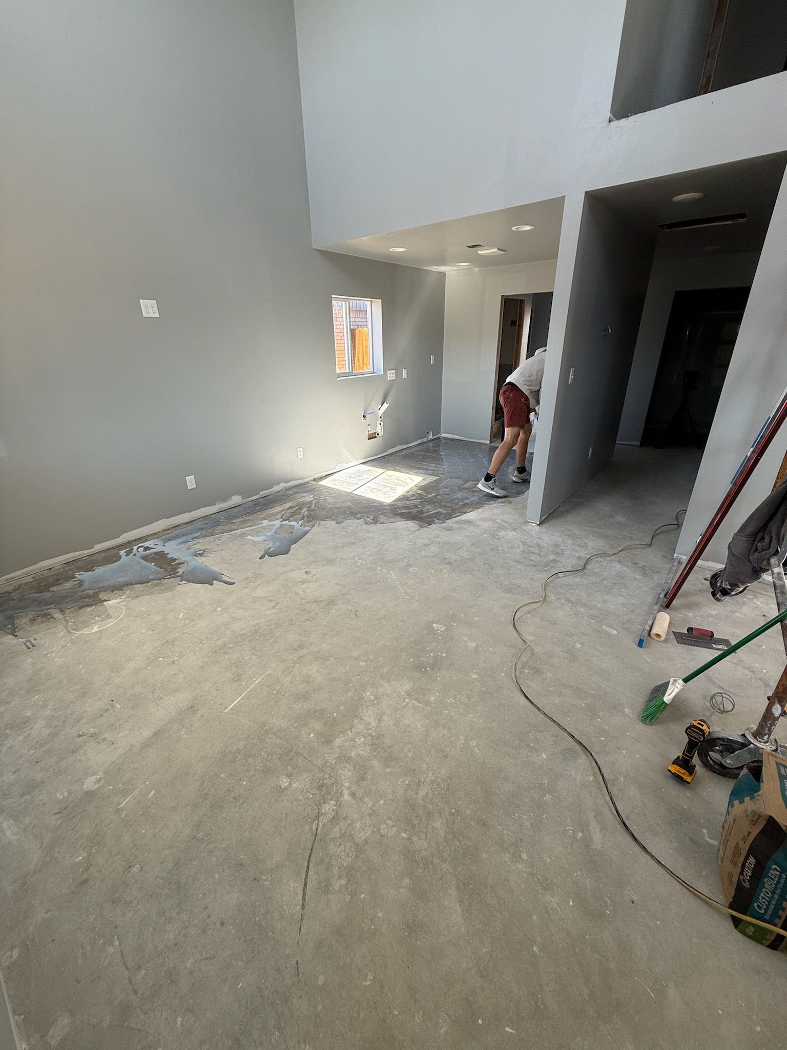 Person applying flooring in a room with gray walls. Concrete floor, tools visible.