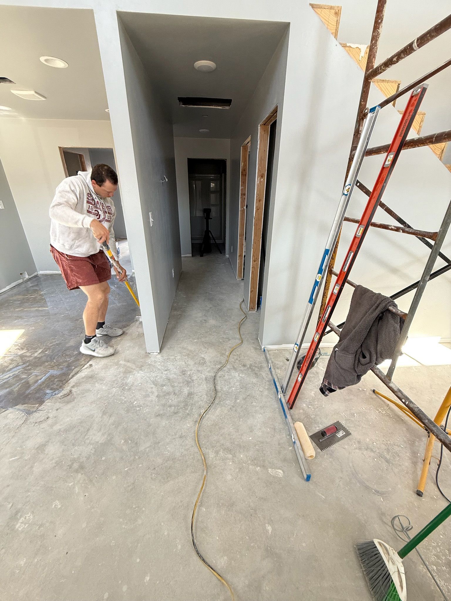 Person sweeping debris in a construction area. Hallway, gray walls, ladder, and tools visible.
