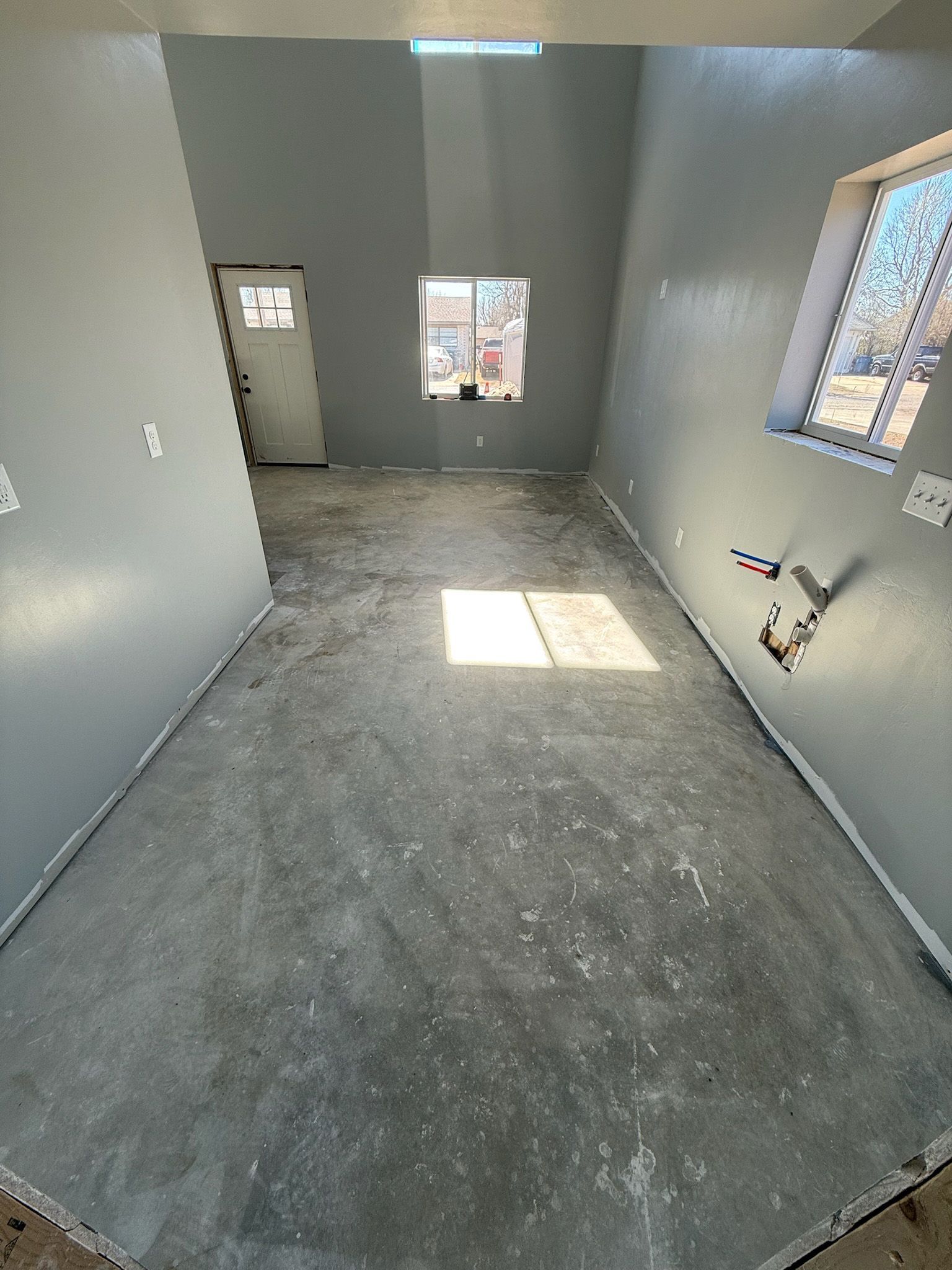 Empty room with gray walls, concrete floor, a door, and windows, appearing under construction.