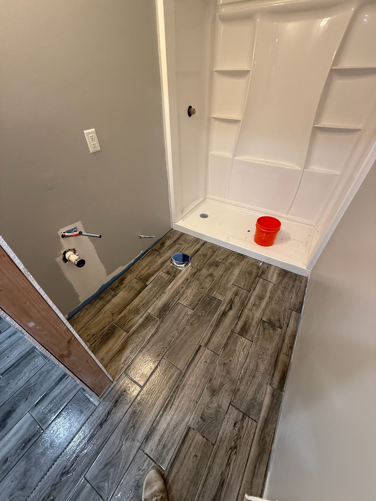 Bathroom renovation in progress. Gray wood-look tile floor, white shower stall, red bucket, gray wall, and unfinished doorway.