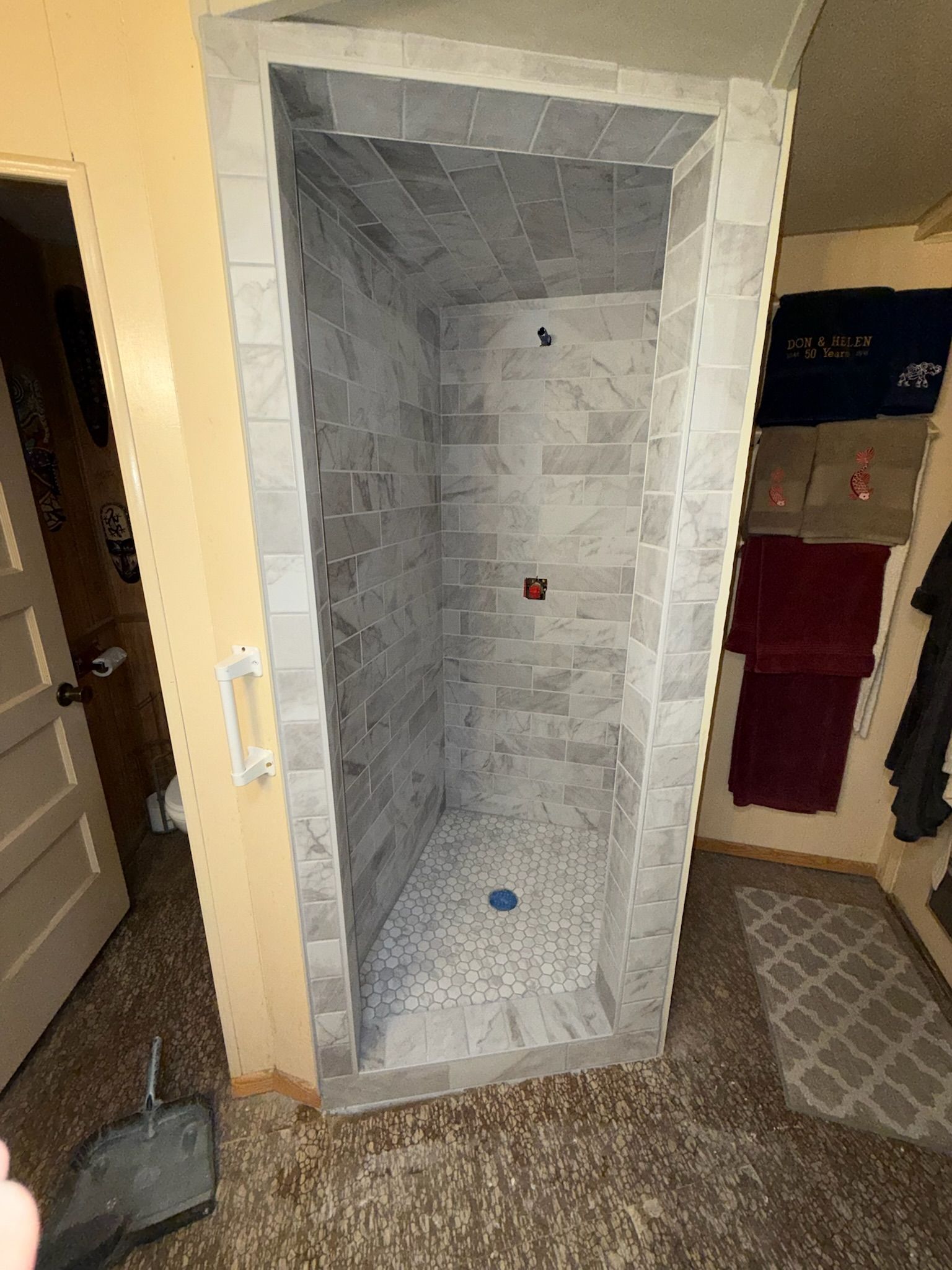 Empty tiled shower stall with gray brick-like tiles, and a mosaic floor, adjacent to a bathroom.
