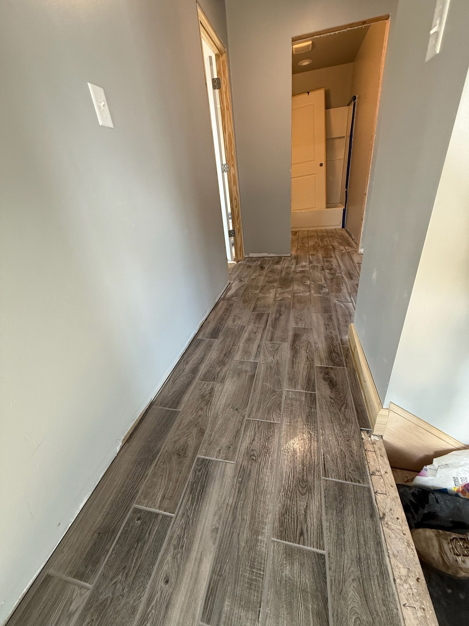 A narrow hallway with gray wood-look flooring and walls. An open doorway leads to another room.