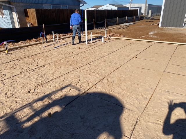 Construction site: Man stands near concrete patio being prepared for pouring. Rebar grid, pipes visible. Sunny day.
