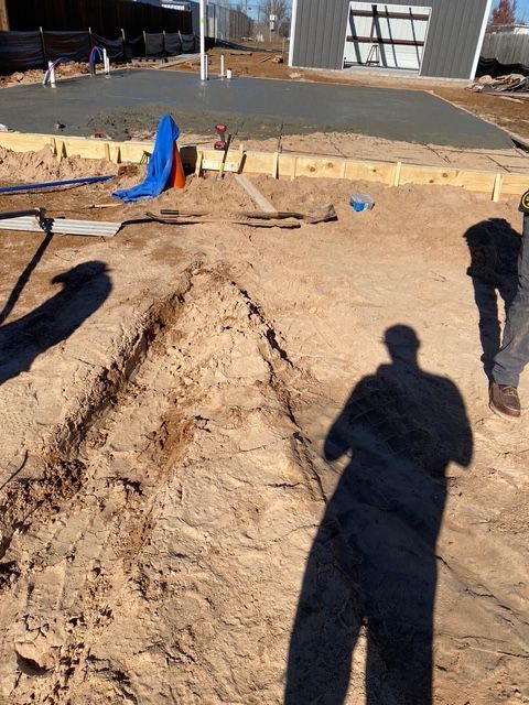Construction site: trench in dirt, freshly poured concrete slab, worker, and building in the background.