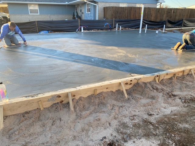 Two workers smoothing fresh concrete slab at a construction site, sunny outdoor setting.