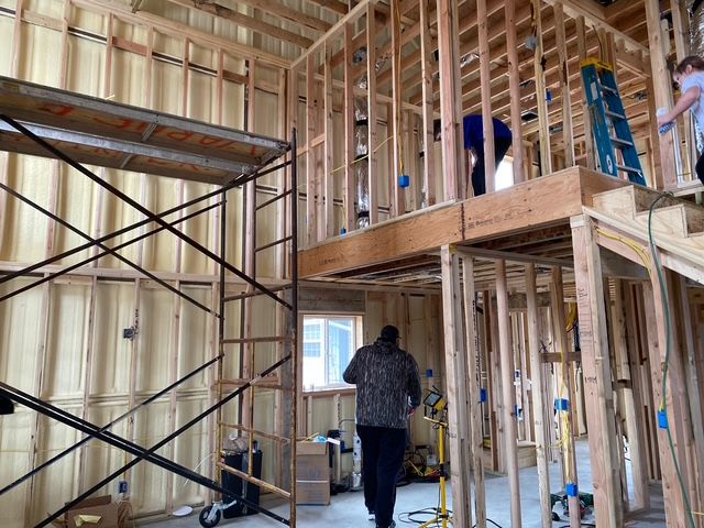 Interior view of a house under construction; wooden framing, insulation, scaffolding, and people working on a mezzanine.