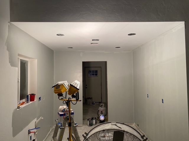 Interior room being painted, lit by work lights. Grey walls, recessed ceiling lights, ladder.