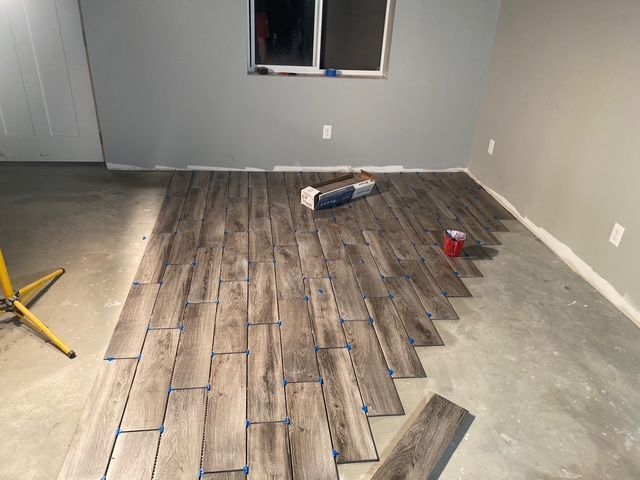 Floor tiling in progress; gray wood-look tiles on a concrete floor, with tools and supplies in a room.