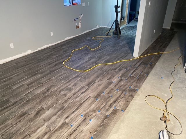 Basement floor installation in progress; gray wood-look tile, yellow extension cord.