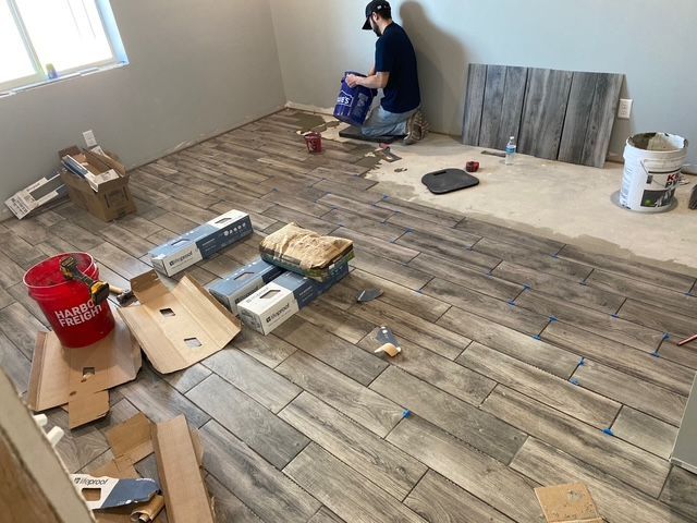 Man tiling a room with wood-look tiles; supplies and boxes are scattered around the work in progress.