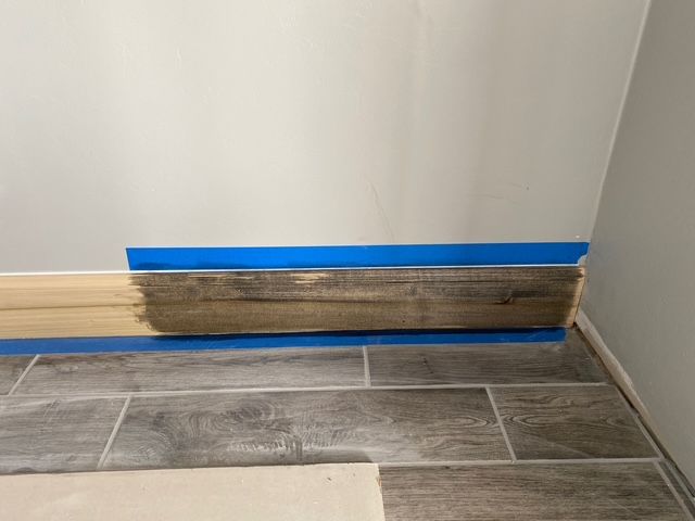 Baseboard being stained; blue tape protects wall and floor tiles.
