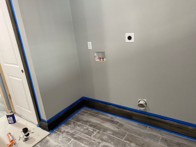 Laundry room corner with gray walls, a white door, and blue painter's tape on trim, floor, and a power outlet.