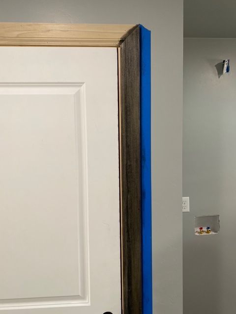 Close-up of a white door with stained dark wood trim and blue painter's tape. Light-colored trim above.