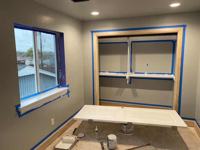 Room with window and closet being painted. Blue tape protects surfaces, white paint, neutral walls.