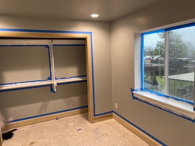 Room with freshly painted light gray walls, blue tape around a closet and window, and wood trim.