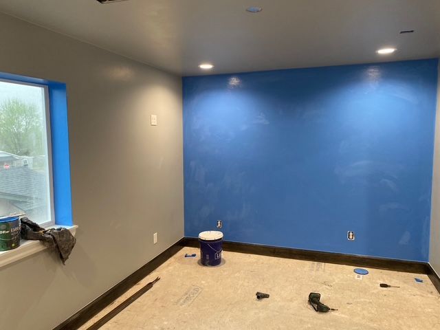 Room with gray and blue walls, construction in progress. Taped window, paint bucket, and tools on the floor.