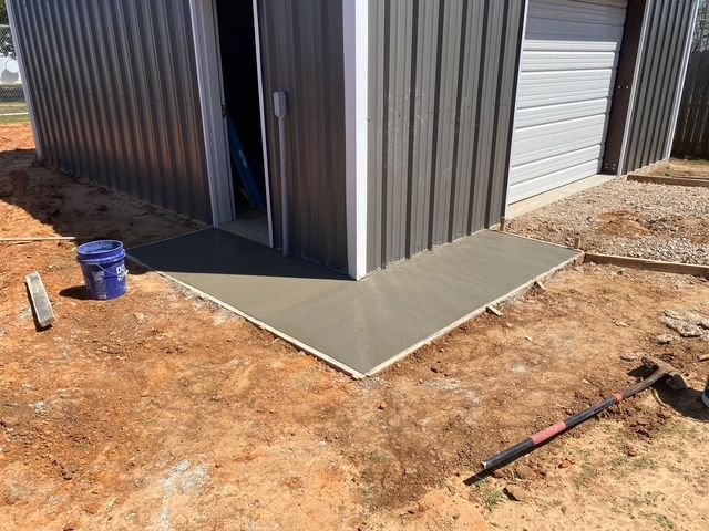 Freshly poured concrete ramp at corner of metal building. Brown soil, blue bucket, tools.
