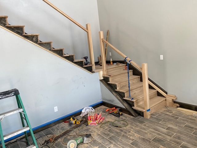 Staircase construction with wood steps, railing, and tools, against gray walls and tiled floor.