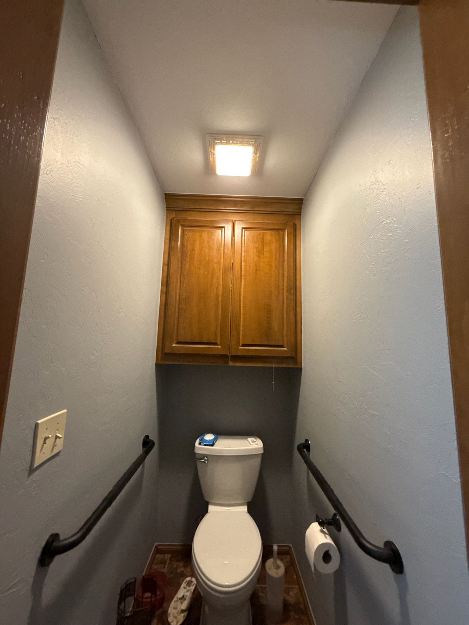 Small bathroom with toilet, wood cabinet, grab bars, and overhead light.