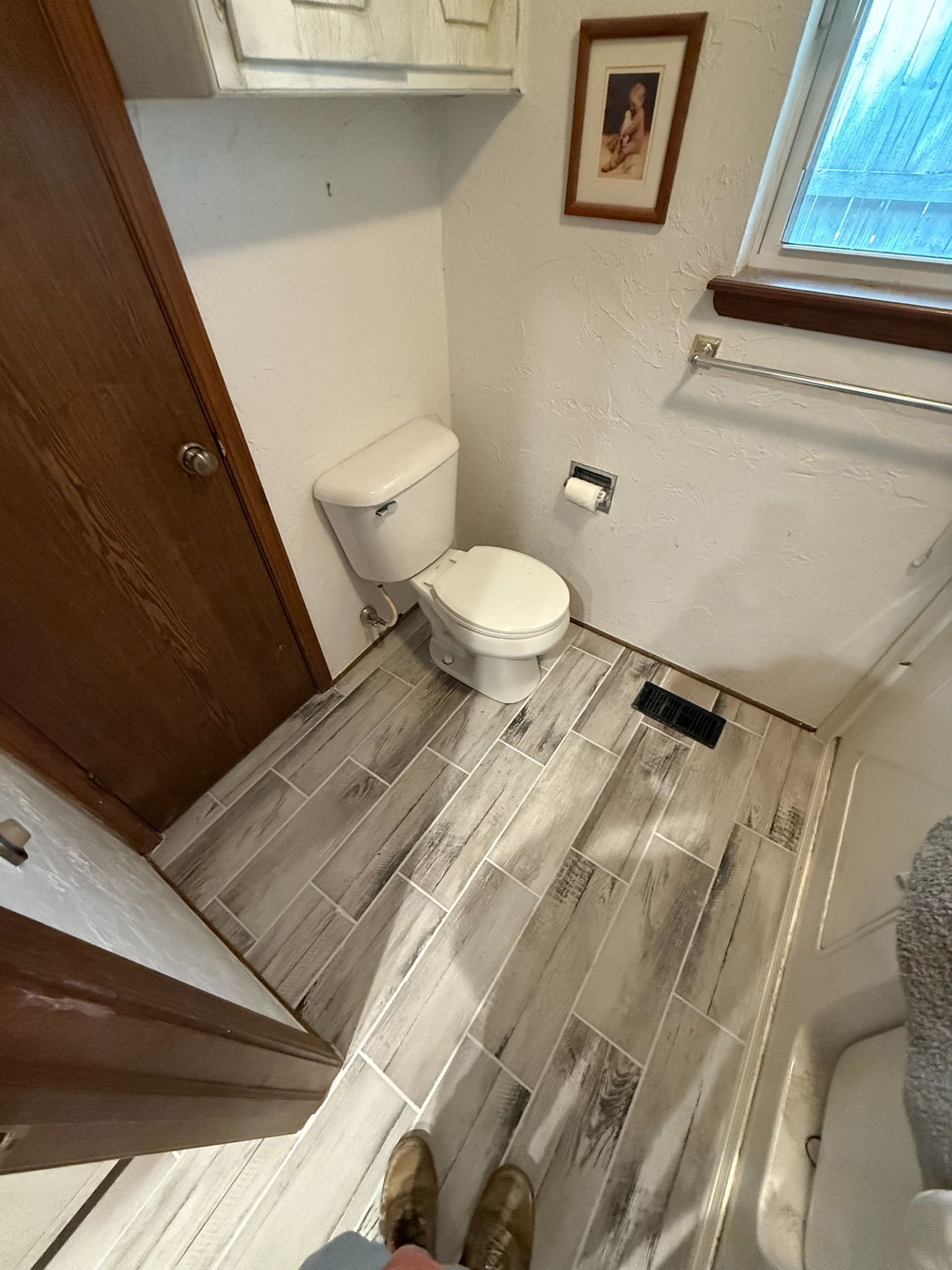 Small bathroom with toilet, new flooring, and a window. Brown door on the left.