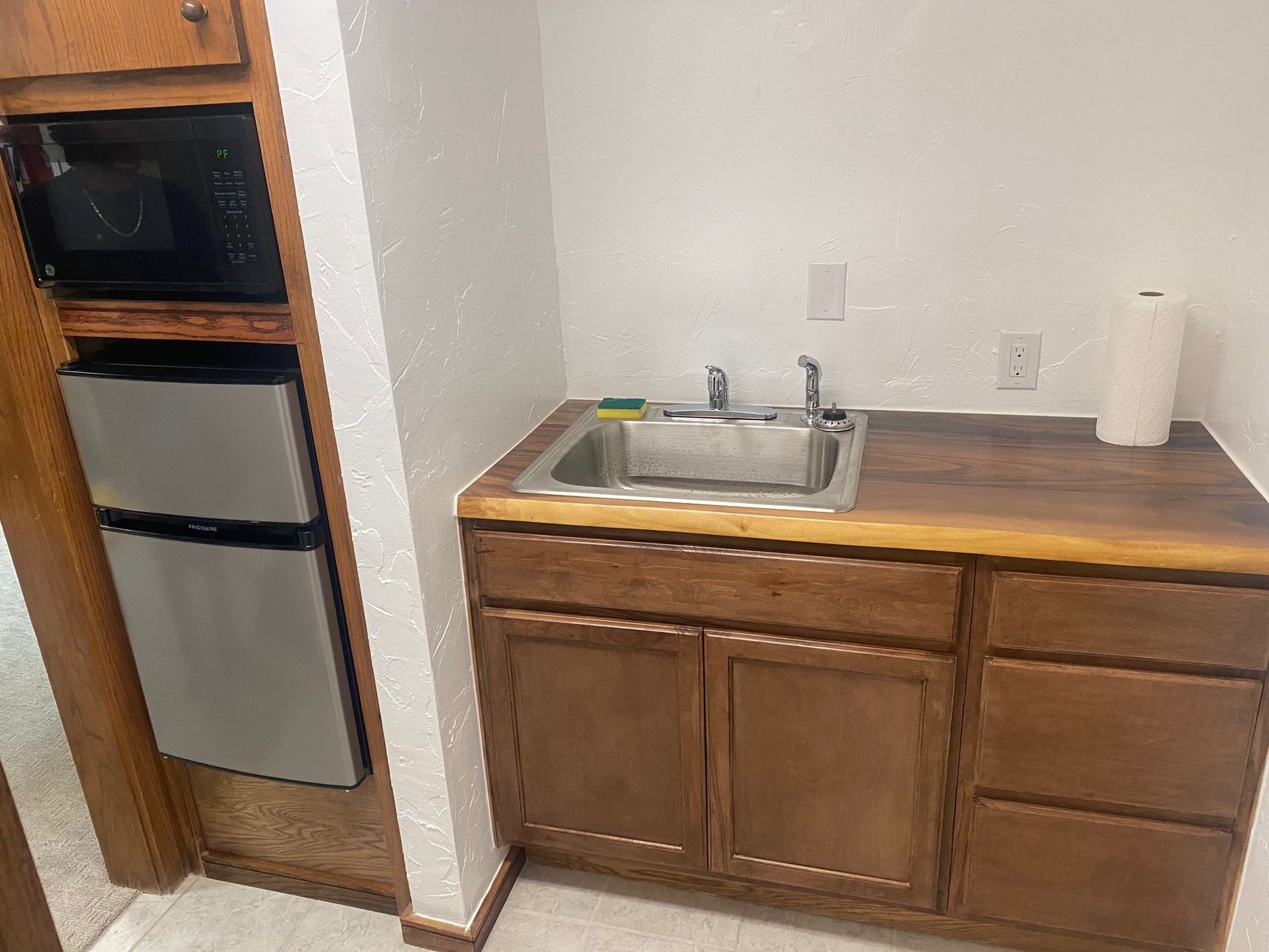 Small kitchen with a sink, cabinets, microwave, and mini-fridge. Brown wood and neutral tones.