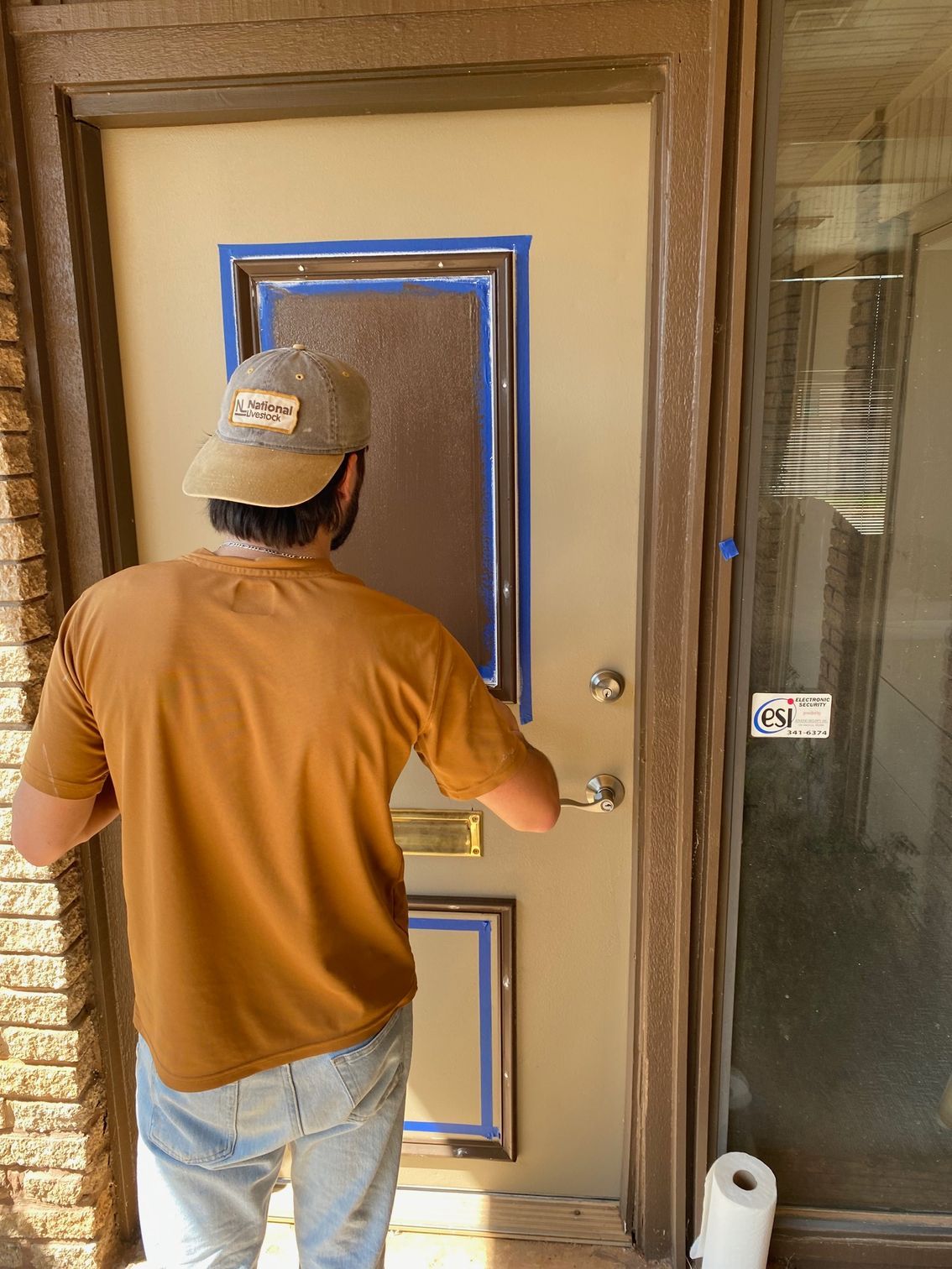 Person taping a door for painting; tan shirt, hat, blue tape, beige door, brown frame.