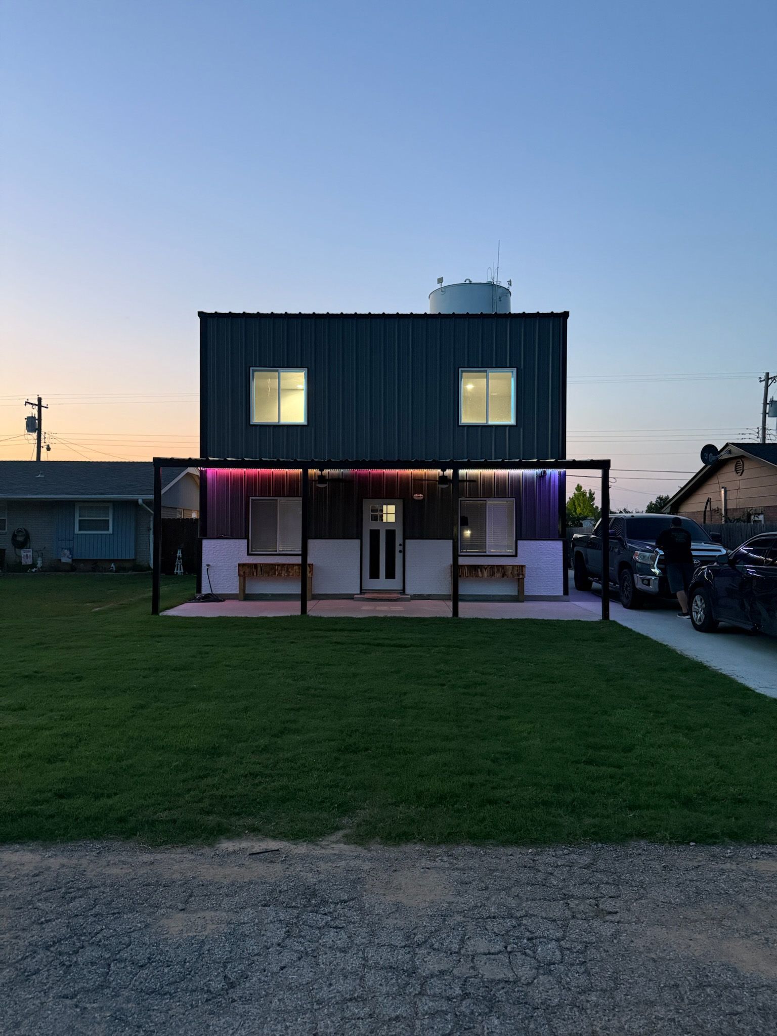 Two-story building made from shipping containers with porch and colorful lights at dusk.