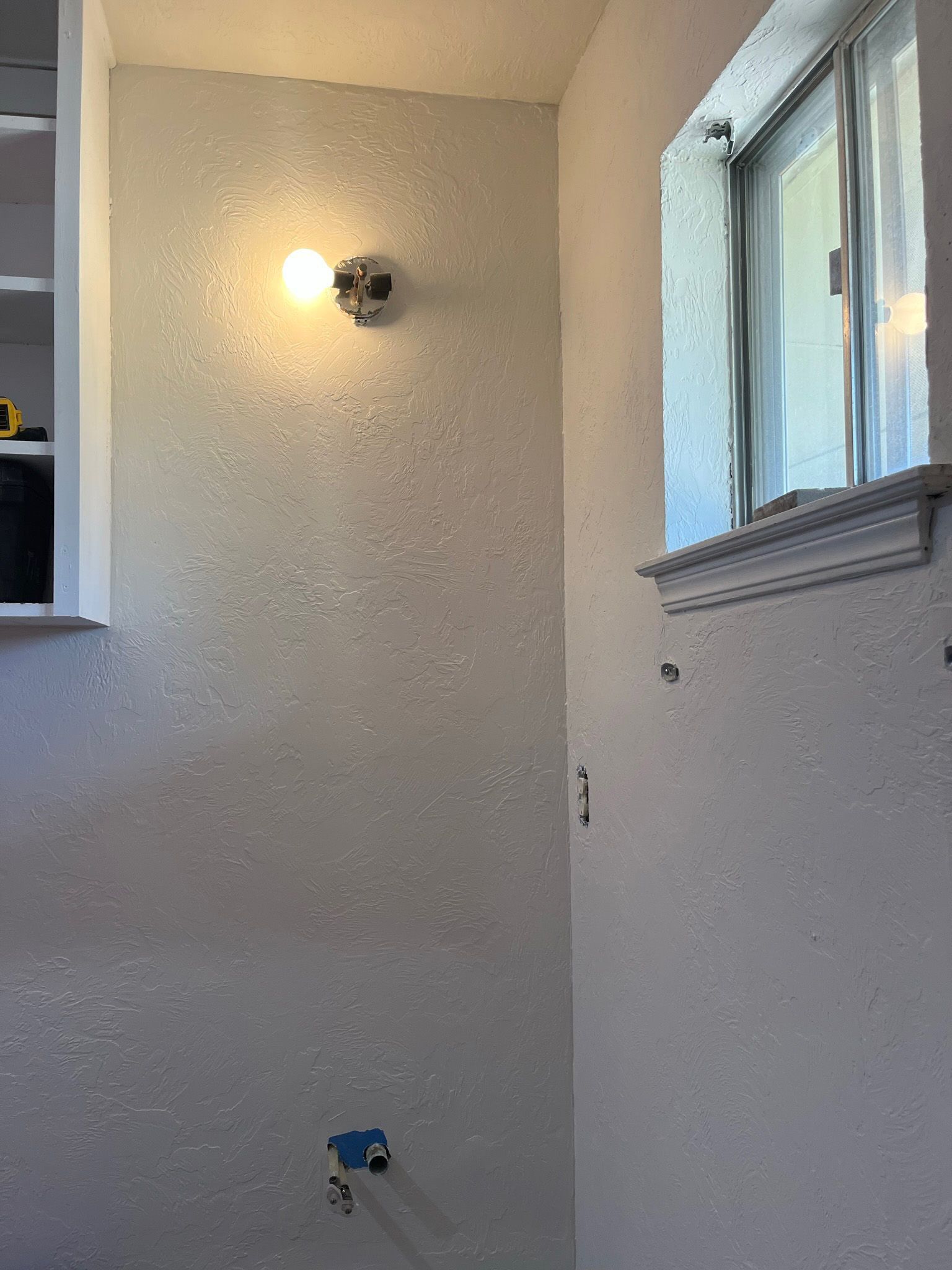 White-walled corner with a window, light fixture, and exposed plumbing.